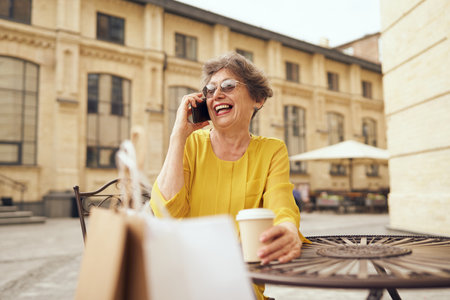 Happy mature woman sitting at cafe terrace, drinking coffee and having telephone conversationの写真素材