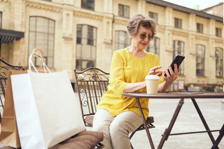 Smiling senior woman is using smartphone while sitting at cafe terrace with coffee after shoppingの写真素材