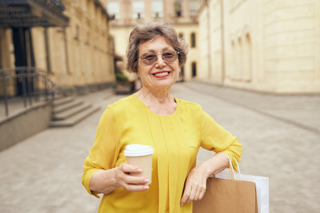 Happy mature woman walking with her shopping purchases and coffee cup in city streetの写真素材