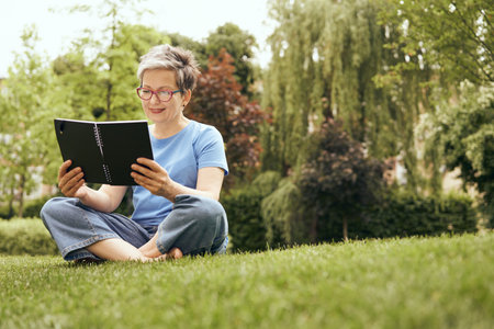 Senior woman in eyeglasses reading book while sitting on grass in park and having a restの写真素材