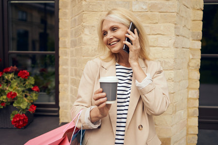 Close up of mature woman with shopping bags standing in city while drinking coffee and talking phoneの写真素材