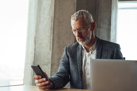 Focused senior executive ceo looking on smartphone during work on laptop in officeの写真素材