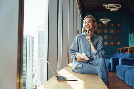 Smiling young woman freelancer sitting near window and making notes during working on laptopの写真素材