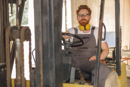 Forklift driver in protective eyeglasses sitting in vehicle in wood warehouse and looks camera