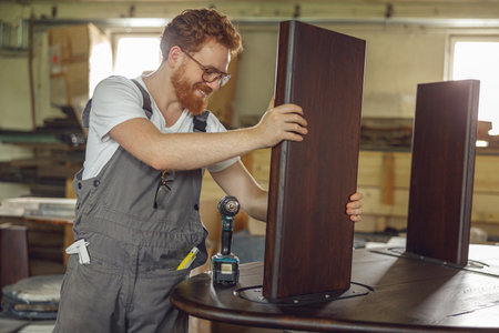 Smiling carpenter fixing table with electric drill standing in carpentry workshopの写真素材