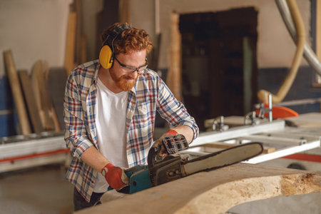 Woodworker in glasses is cutting wood with chainsaw on carpentry workshop. High quality photoの写真素材
