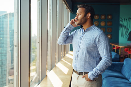 Focused entrepreneur is talking phone with client while standing in modern coworking near windowの写真素材