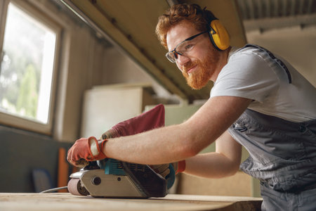 Bearded male wood worker using electric sander for wood. Carpentry workshop. High quality photoの写真素材