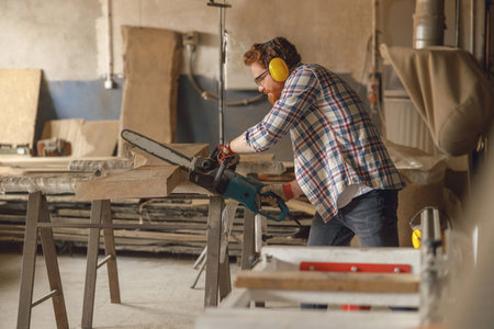 Professional carpenter in glasses is cutting wood with chainsaw on carpentry workshopの写真素材