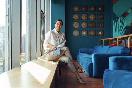 Attractive woman office worker holding notepad sitting in coworking and looking at cameraの写真素材