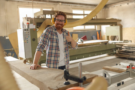 Smiling carpenter cutting wooden board at sawmill carpentry manufacturing and showing thumb upの写真素材