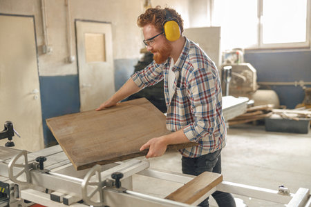 Smiling carpenter putting wooden board on circular saw at carpentry manufacturingの写真素材