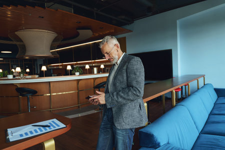 Senior businessman is holding phone while standing in office during working dayの写真素材