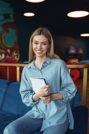 Pretty woman freelancer holding notepad and looking at camera with smile in coworkingの写真素材