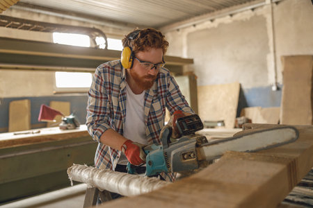 Professional carpenter in glasses is cutting wood with chainsaw on carpentry workshopの写真素材