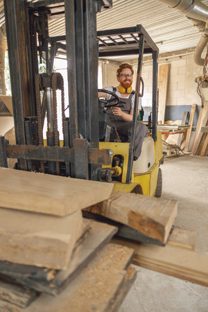 Male carpenter at wooden sawmill warehouse is loading with loader wooden board