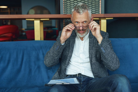 Portrait of senior businessman analyst in eyeglasses sitting on sofa in office and looking at sideの写真素材