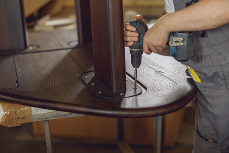 Close up of carpenter fixing table with electric drill standing in carpentry workshop and looks awayの写真素材