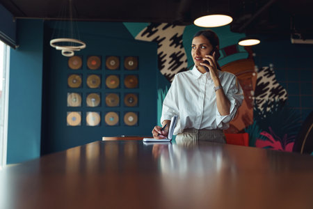 Stylish busy female entrepreneur making notes during phone conversation with client in coworkingの写真素材