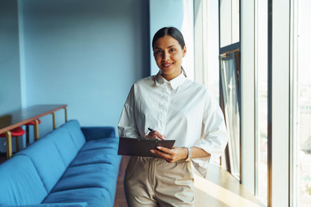 Smiling female sales manager holding clipboard while standing in office near windowの写真素材