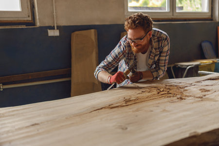 Carpenter in glasses working with chisel while cutting wooden plank in carpentry workshopの写真素材