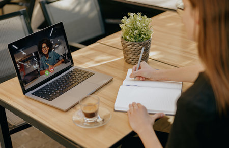 Businesswoman talking with client via video call and making notes while sitting in cafe terrace