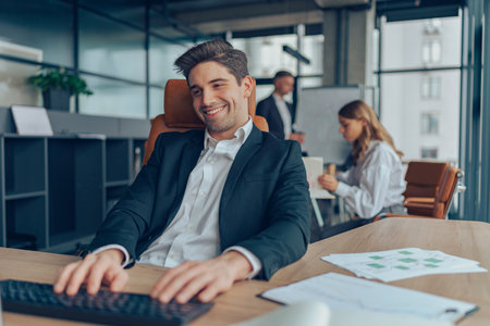 Smiling handsome male manager working on computer while sitting in office on colleagues backgroundの写真素材