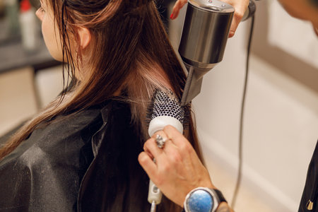 Close up of professional female barber with hair dryer and brush styling hair of customer in salonの写真素材