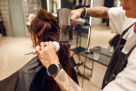 Close up of professional female barber with hair dryer and brush styling hair of customer in salonの写真素材