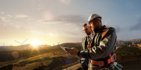 Windmill engineer and businessman planing new ecology project. they standing and looking in tablet. Around wind generators blueprint and beautiful landscapeの写真素材