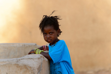 FERLO DESERT, SENEGAL - APR 25, 2017: Unidentified Fulani little girl in blue suit leans on the water-draw. Fulanis (Peul) are the largest tribe in West African savannahsのeditorial素材
