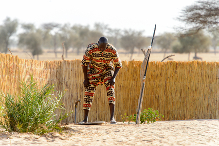 FERLO DESERT, SENEGAL - APR 25, 2017: Unidentified Fulani man in colored suit bends down to the water taps . Fulanis (Peul) are the largest tribe in West African savannahsのeditorial素材