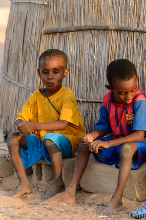 FERLO DESERT, SENEGAL - APR 25, 2017: Unidentified Fulani little boys sit on bricks near the fence. Fulanis (Peul) are the largest tribe in West African savannahsのeditorial素材