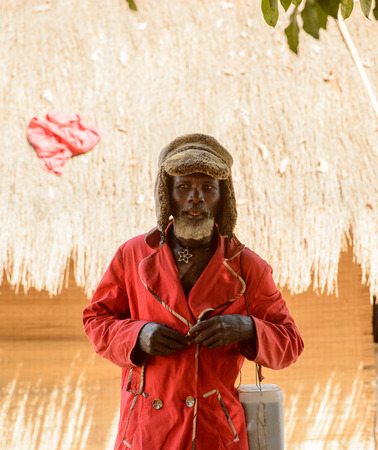 CANHABAQUE, GUINEA BISSAU - MAY 4, 2017: Unidentified local man with beard in a hat and red coat stands near the shack in a village of the Canhabaque island. People in G.-Bissau still suffer of povertyのeditorial素材