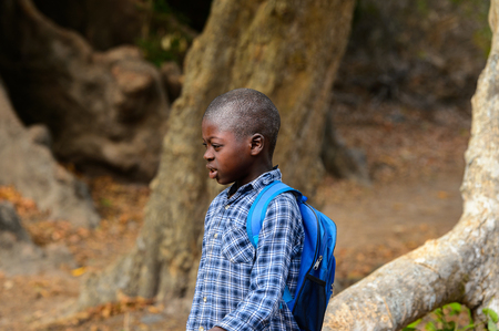 SOGA, GUINEA BISSAU - MAY 5, 2017: Unidentified local little boy in plaid shirt with backpack walks along the street in a village of the Soga island. People in G.-Bissau still suffer of povertyのeditorial素材