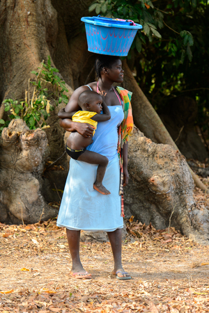 SOGA, GUINEA BISSAU - MAY 5, 2017: Unidentified local woman carries a basin on her head and a little baby in her hand in a village of the Soga island. People in G.-Bissau still suffer of povertyのeditorial素材
