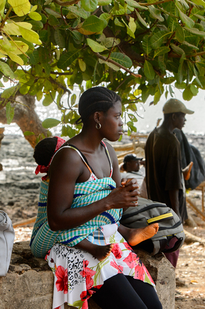 SOGA, GUINEA BISSAU - MAY 5, 2017: Unidentified local woman holds a little baby on her back in a village of the Soga island. People in G.-Bissau still suffer of povertyのeditorial素材