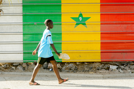 DAKAR, SENEGAL - APR 23, 2017: Unidentified Senegalese boy walks with a pair of shoes in front of...