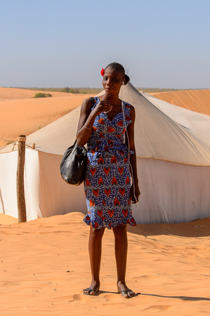 LAMPOUL DESERT, SENEGAL - APR 23, 2017: Unidentified Senegalese woman with braids wears red stripe on her head and colored dress holds a bag in a Lampoul Desert, beautiful landscape placeのeditorial素材