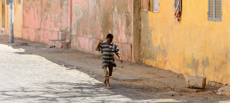 SAINT LOUIS, SENEGAL - APR 24, 2017: Unidentified Senegalese little boy in striped shirt walks with a bag along the road near the building in Saint Louis, one of the biggest cities in Senegalのeditorial素材