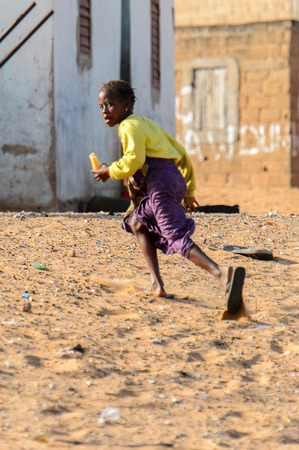 KAYAR, SENEGAL - APR 27, 2017: Unidentified Senegalese little girl with braids runs along the road with a piece of bread in a beautiful village near Kayar, Senegalのeditorial素材