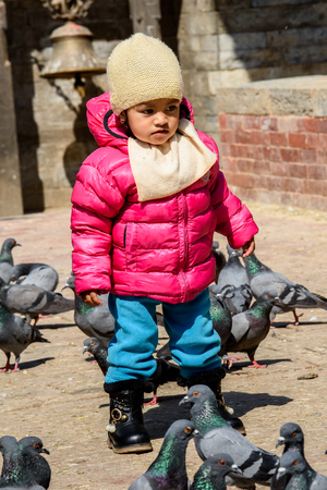 KATMANDU, NEPAL - MAR 6, 2017: Unidentified Chhetri little girl in pink jacket and a hat walks around pigeons. Chhetris is the most populous ethnic group of Nepalのeditorial素材