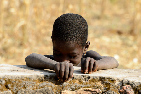 KOUTAMMAKOU, TOGO - JAN 13, 2017: Unidentified Togolese little girl stands behind the rock in the village. Togo children suffer of poverty due to the bad economy.のeditorial素材