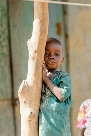 GHANI, GHANA - JAN 14, 2017: Unidentified Ghanaian little boy in green shirt leans on the wooden tree in the Ghani village. Ghana children suffer of poverty due to the bad economy.のeditorial素材