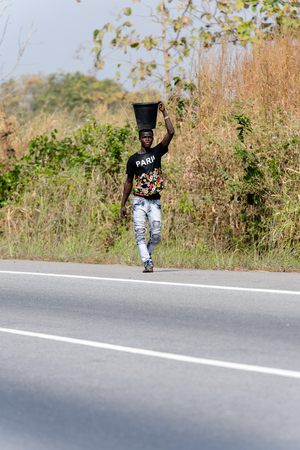 BRONG AHAFO, GHANA - JAN 15, 2017: Unidentified Ghanaian man in black shirt carries a bucket on his head. Ghana people suffer of poverty due to the bad economy.のeditorial素材
