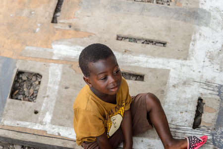 ACCRA, GHANA - JAN 8, 2017: Unidentified Ghanaian little boy in mustard shirt sits on the wooden board. Children of Ghana suffer of poverty due to the economic situationのeditorial素材