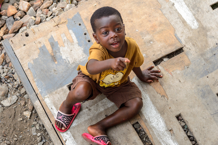 ACCRA, GHANA - JAN 8, 2017: Unidentified Ghanaian little boy in mustard shirt sits on the wooden board. Children of Ghana suffer of poverty due to the economic situationのeditorial素材
