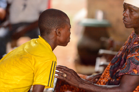 LOME, TOGO - Jan 9, 2017: Unidentified Togolese young boy in yellow shirt bends to the local shaman in his house. Togo people suffer of poverty due to the bad economyのeditorial素材