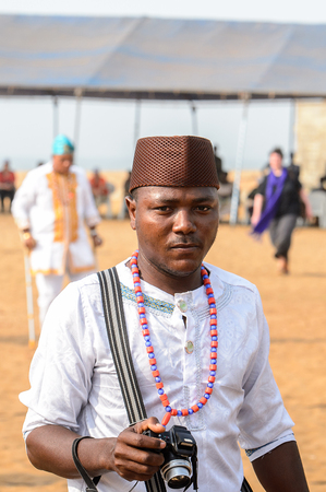 OUIDAH, BENIN - Jan 10, 2017: Unidentified Beninese man in white shirt and a hat wears necklace and carries a camera at the voodoo festival, which is anually celebrated on January, 10th.のeditorial素材