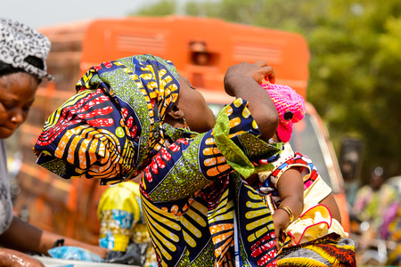 OUIDAH, BENIN - Jan 10, 2017: Unidentified Beninese woman in colored clothes carries her baby at the local market. Benin people suffer of poverty due to the bad economyのeditorial素材